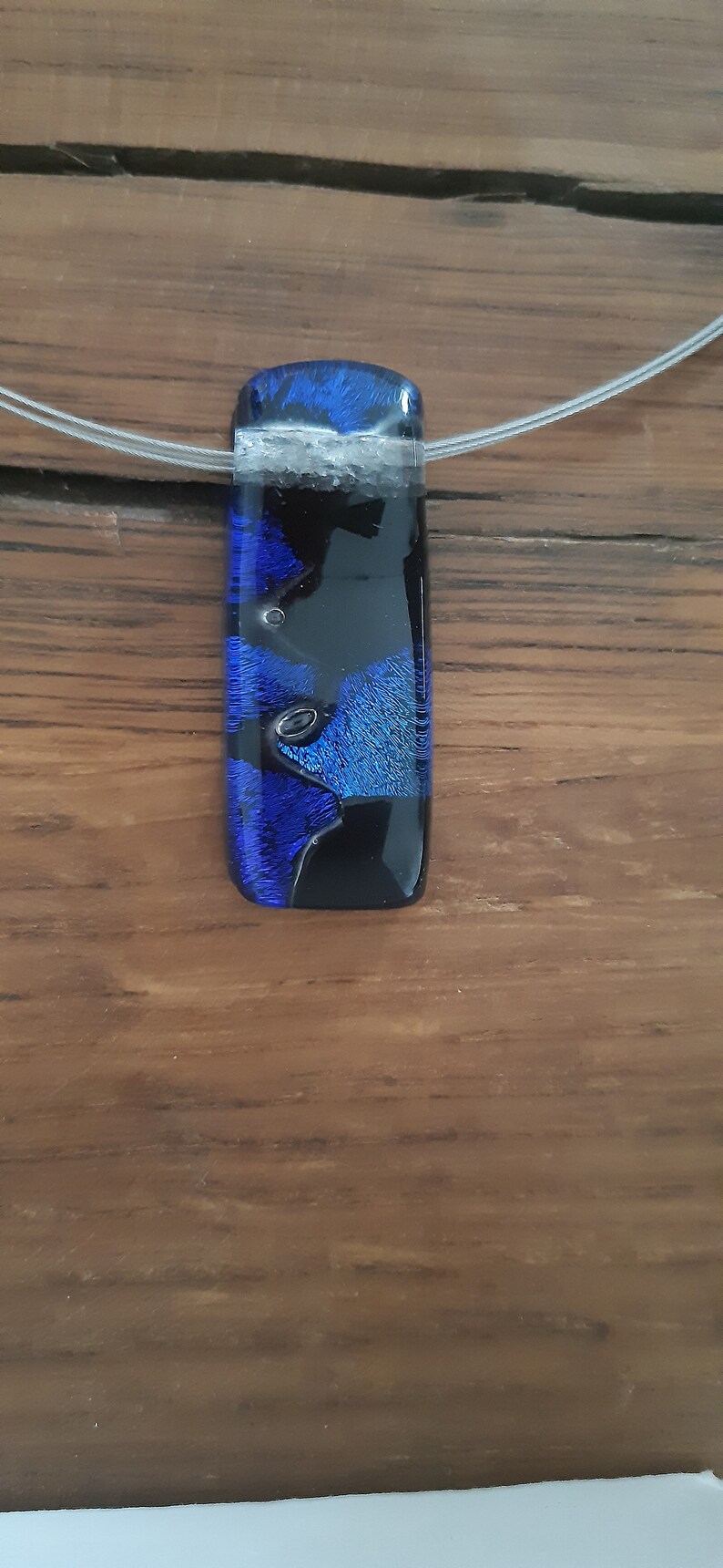 K&ouml;nnte beinhalten: Ein blauer und schwarzer Glasanh&auml;nger mit einer silbernen Kette. Der Anh&auml;nger hat ein einzigartiges, abstraktes Design mit Wirbeln aus blauem und schwarzem Glas.
