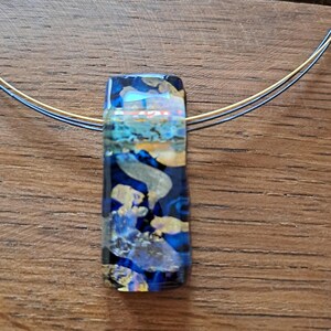 May include: A rectangular pendant necklace with a blue, gold, and teal design. The pendant is suspended from a thin, gold-colored chain. The pendant's design features layers of color and texture, creating a unique and artistic look. The necklace is displayed on a wooden surface.