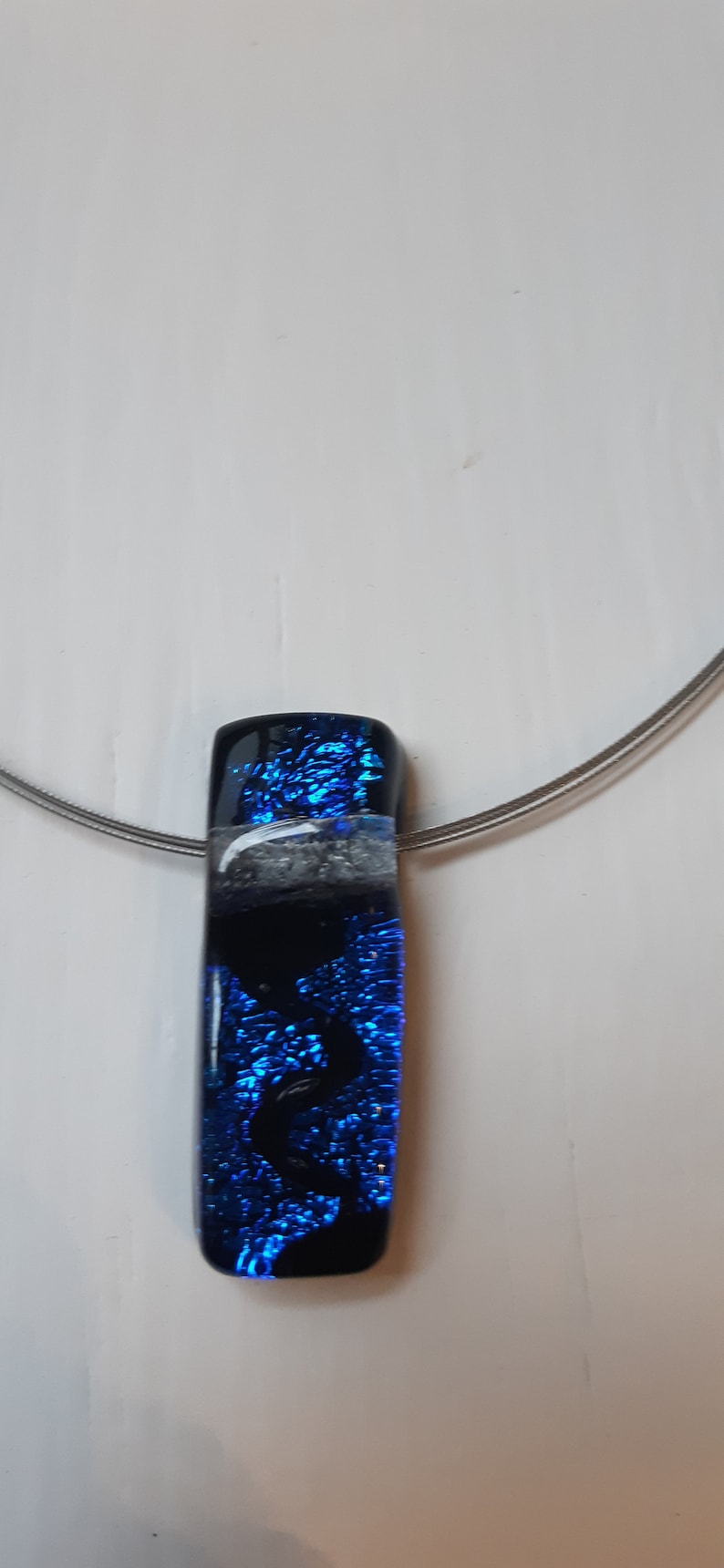 K&ouml;nnte beinhalten: Eine silberne Kettenkette mit einem blauen und schwarzen Glasanh&auml;nger. Der Anh&auml;nger hat einen wei&szlig;en Streifen und eine glitzernde blaue Oberfl&auml;che.