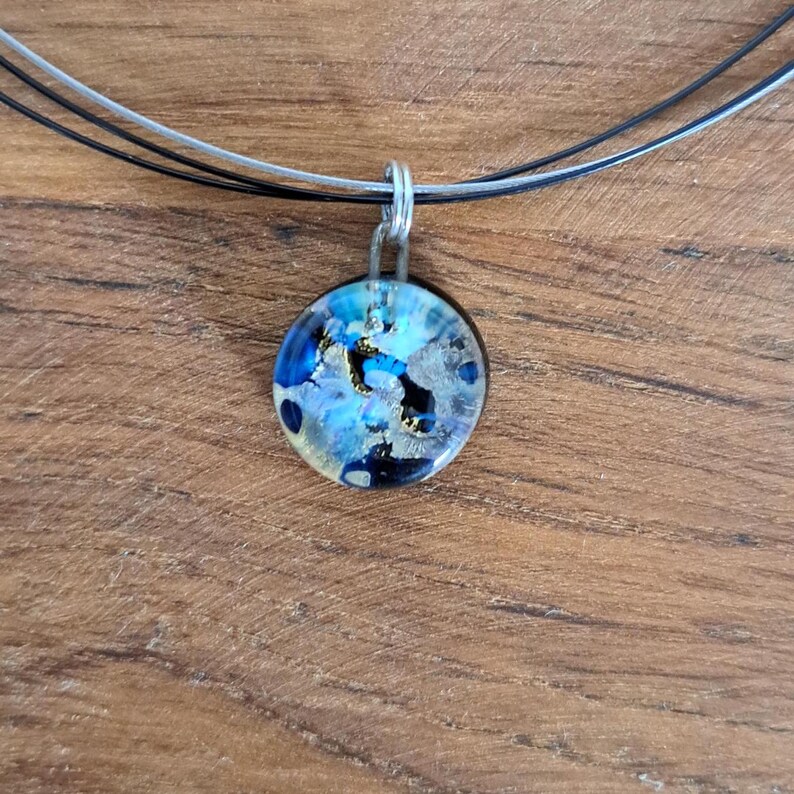 Puede incluir: Un collar de cadena de plata con un colgante redondo de vidrio. El colgante tiene un dise&ntilde;o abstracto azul, blanco y dorado.