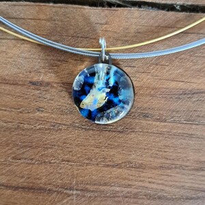 Puede incluir: Un collar de plata con un colgante redondo de vidrio azul y dorado. El colgante tiene un patr&oacute;n de remolino de azul, dorado y blanco.