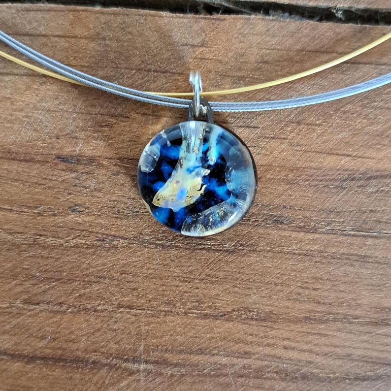 Puede incluir: Un collar de plata con un colgante redondo de vidrio azul y dorado. El colgante tiene un patr&oacute;n de remolino de azul, dorado y blanco.