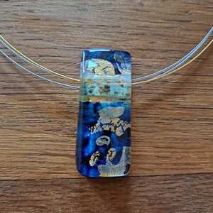 May include: A rectangular pendant necklace with a blue, gold, and silver design. The pendant has a layered appearance with gold foil accents and is suspended from a silver and gold-tone wire necklace.