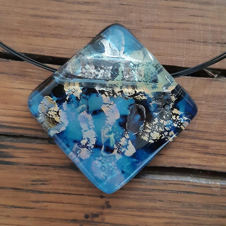 May include: A square-shaped glass pendant necklace with a blue, black, and gold abstract design. The pendant is on a black cord.