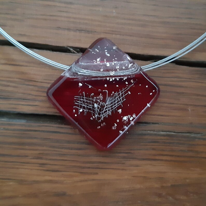 May include: A red glass pendant with a white design inside. The pendant is shaped like a diamond and is hanging from a silver chain.