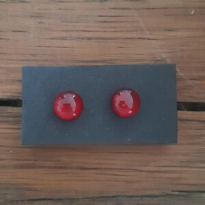May include: A pair of red, round stud earrings. The earrings are set on a small, rectangular black display card. The background is a wooden surface.