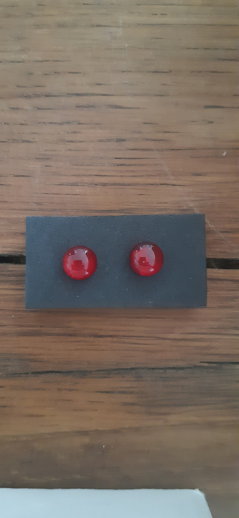 May include: A pair of red, round stud earrings. The earrings are set on a small, rectangular black display card. The background is a wooden surface.