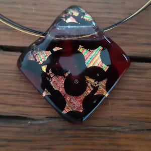 May include: A dark red glass pendant with a black and gold geometric design. The pendant is attached to a thin gold chain.