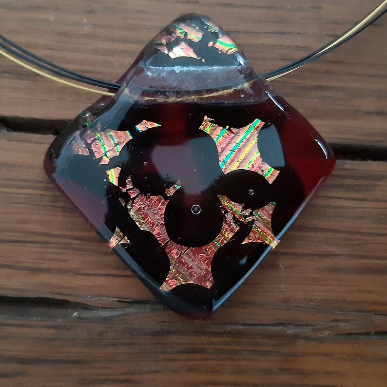May include: A dark red glass pendant with a black and gold geometric design. The pendant is attached to a thin gold chain.