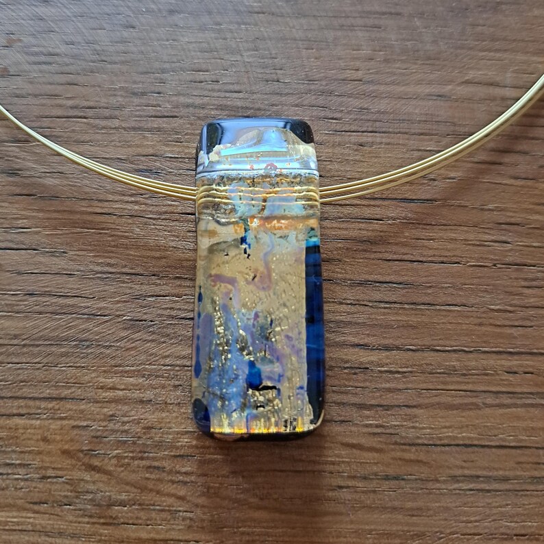 May include: A rectangular pendant necklace with a gold-colored wire choker. The pendant is a mix of blue, gold, and clear glass, with a rectangular shape. The necklace is displayed on a wooden surface.