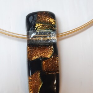 K&ouml;nnte beinhalten: Ein goldener und schwarzer Glasanh&auml;nger mit einer goldenen Kette. Der Anh&auml;nger hat eine strukturierte Oberfl&auml;che und einen klaren Streifen.