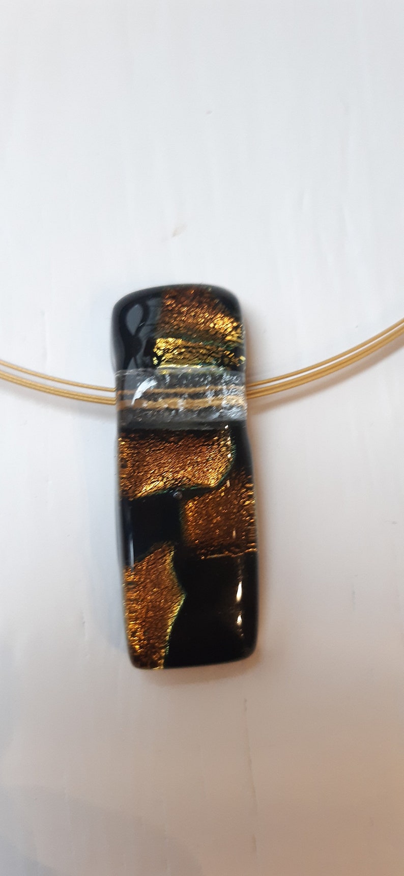 K&ouml;nnte beinhalten: Ein goldener und schwarzer Glasanh&auml;nger mit einer goldenen Kette. Der Anh&auml;nger hat eine strukturierte Oberfl&auml;che und einen klaren Streifen.