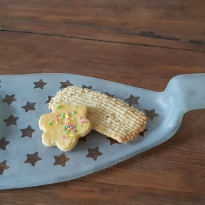 May include: A light blue glass serving tray shaped like a bottle with a star pattern. Two cookies are on the tray, one is a plain cookie and the other is a star-shaped cookie with sprinkles.