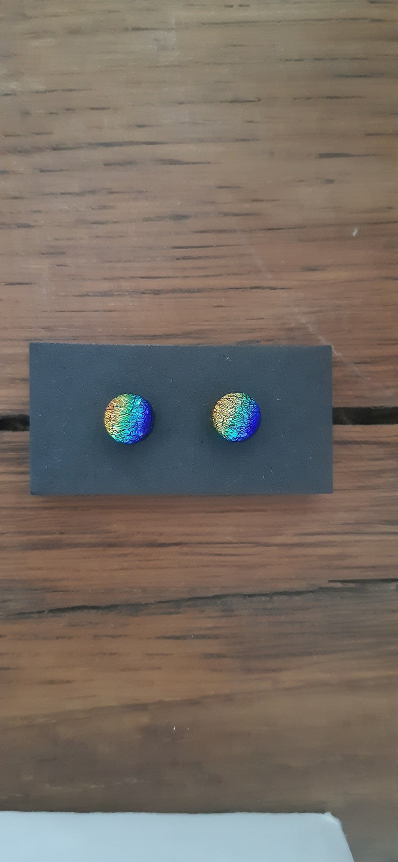 May include: A pair of round, iridescent glass earrings with a yellow, green, and blue color gradient. The earrings are displayed on a black background.