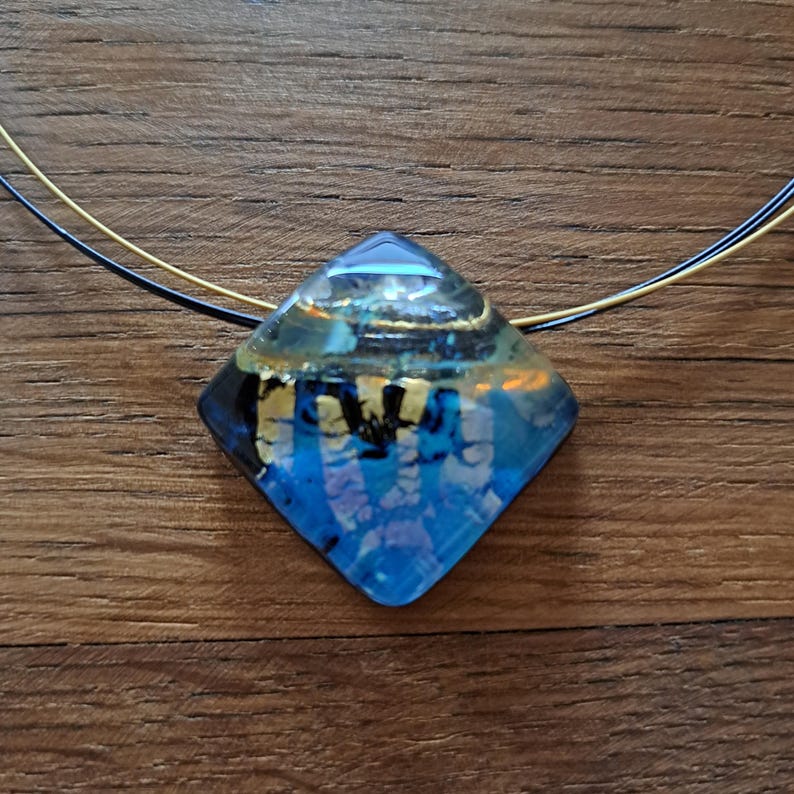 May include: A square-shaped glass pendant necklace with a blue, gold and black abstract design. The necklace is on a black and gold chain.