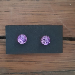 May include: A pair of round, purple dichroic glass stud earrings. The earrings are displayed on a black card against a wooden background. The earrings have a textured, iridescent appearance.