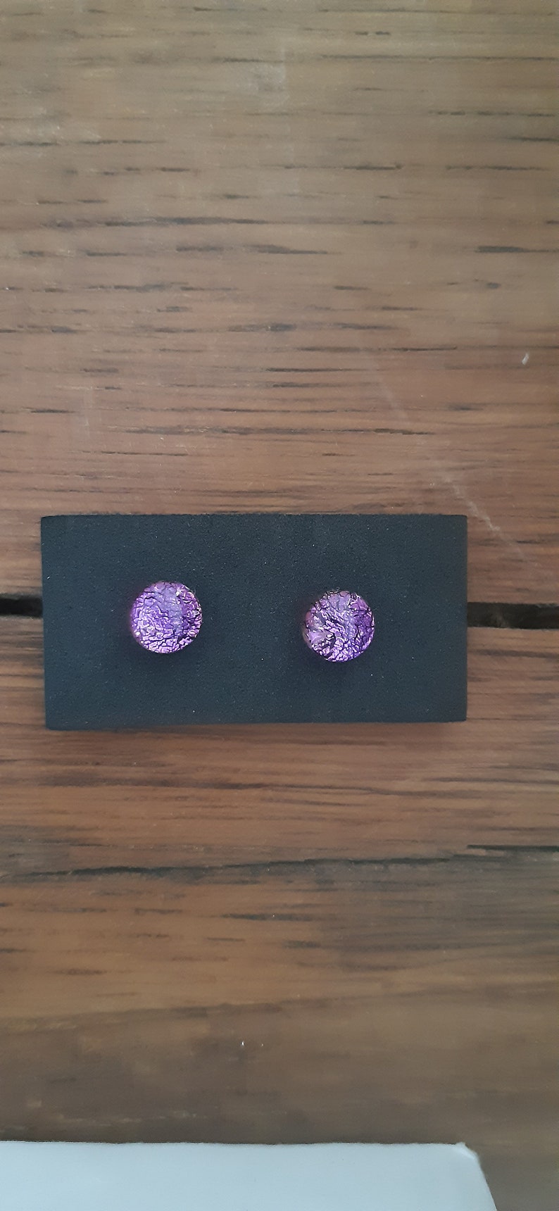 May include: A pair of round, purple dichroic glass stud earrings. The earrings are displayed on a black card against a wooden background. The earrings have a textured, iridescent appearance.