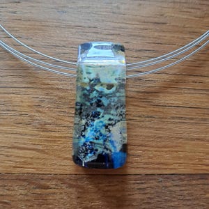 May include: A rectangular pendant necklace with a clear, textured glass design. The pendant features shades of blue, black, and silver, with a clear top. The necklace has a silver-colored wire. The pendant is approximately 1.5 inches long.