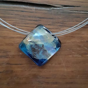 May include: A square-shaped glass pendant necklace with a blue, white and gold marbled design. The pendant is suspended from a silver chain.