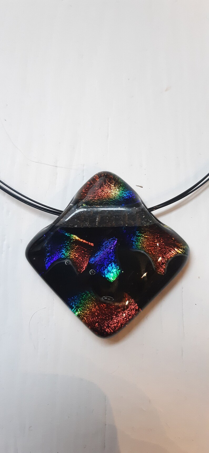 May include: A black glass pendant with iridescent rainbow colors, featuring a textured surface. The pendant is attached to a black cord.