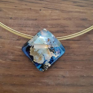 May include: A gold chain necklace with a square-shaped pendant made of blue and gold glass. The pendant has a marbled effect with swirls of blue, gold, and white.