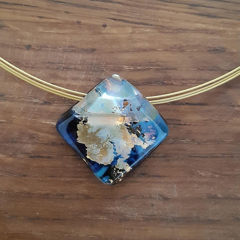 May include: A gold chain necklace with a square-shaped pendant made of blue and gold glass. The pendant has a marbled effect with swirls of blue, gold, and white.