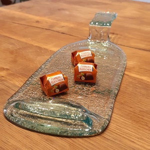 May include: A clear glass bottle shaped into a serving tray with three chocolate candies on top. The candies are wrapped in orange foil and say "Ferrero K&uuml;sschen" on them.
