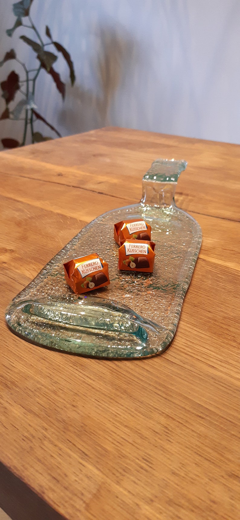 May include: A clear glass bottle shaped into a serving tray with three chocolate candies on top. The candies are wrapped in orange foil and say "Ferrero K&uuml;sschen" on them.