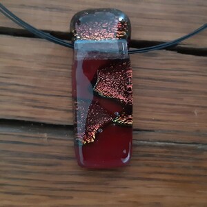 May include: A red and black glass pendant with a textured, iridescent finish. The pendant is rectangular in shape and hangs from a black cord.