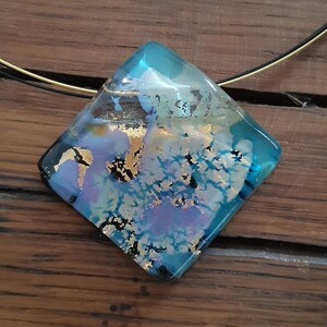 May include: A square-shaped glass pendant necklace with a blue, gold, and purple iridescent design. The pendant is suspended from a thin gold chain.