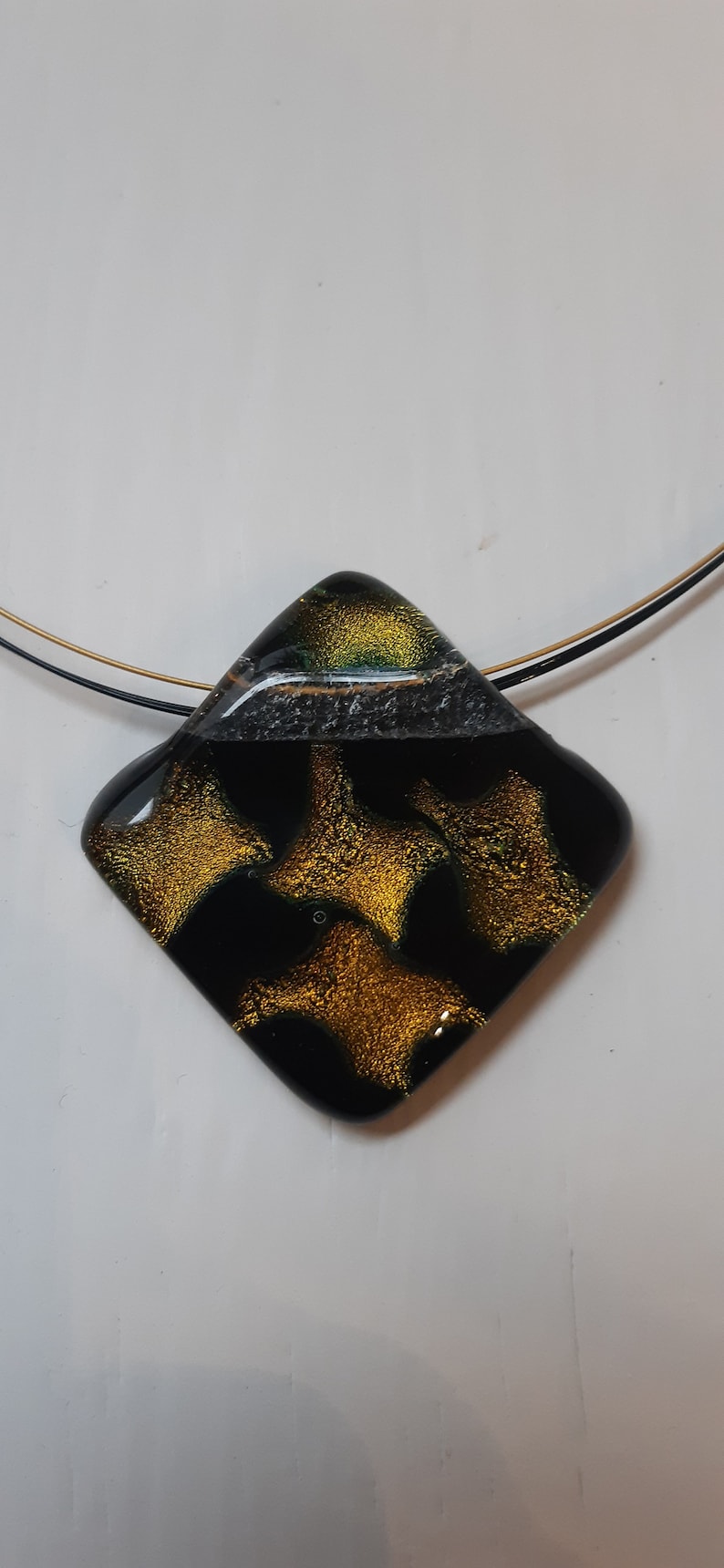 May include: A black and gold glass pendant with a geometric shape and a gold and green pattern. The pendant is hanging from a black and gold chain.