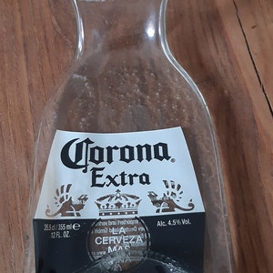 May include: A clear glass bottle shaped like a Corona Extra beer bottle. The bottle has a label with the text "Corona Extra", "35.5 cl/355 ml", "12 FL. OZ.", "Alc. 4.5% Vol.", and "GEM&Auml;SS UNTER LIZENZ VON".
