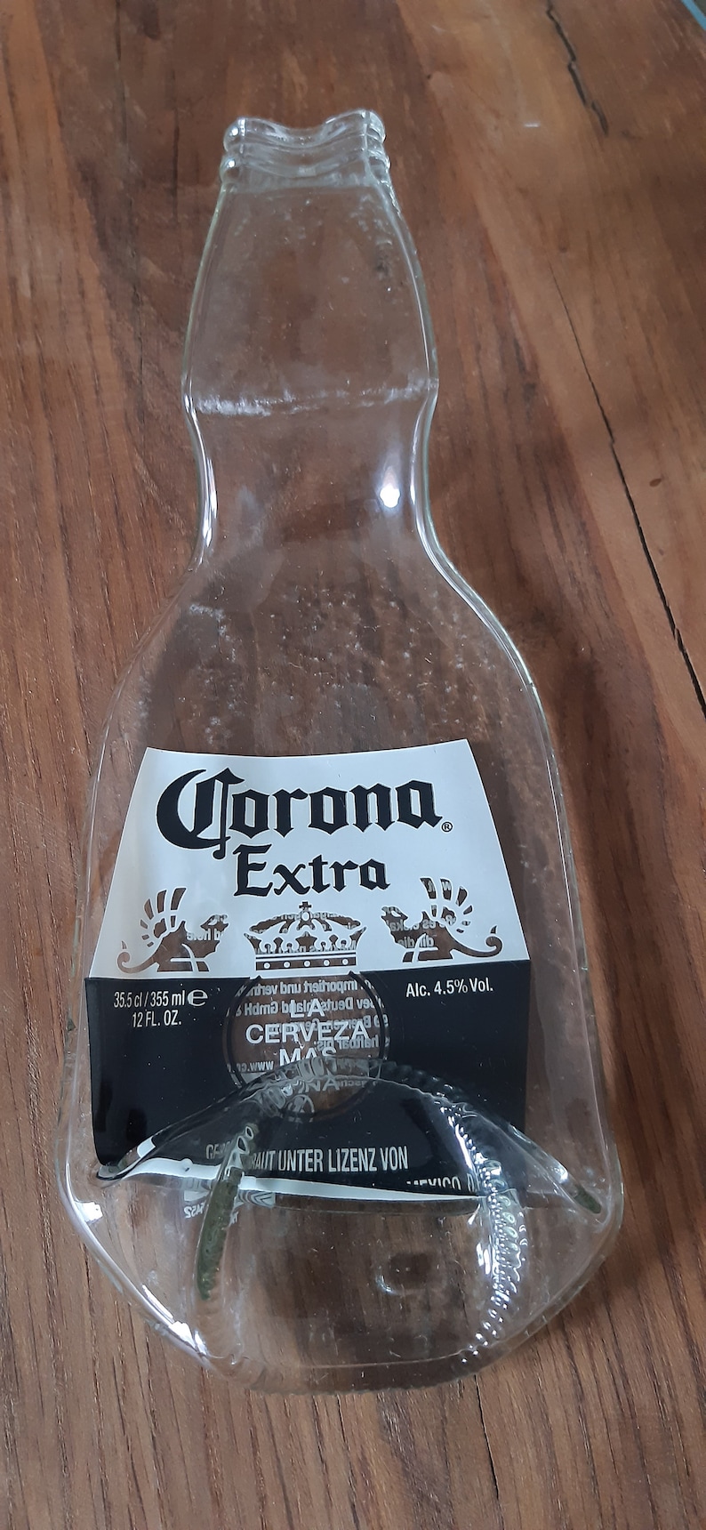May include: A clear glass bottle shaped like a Corona Extra beer bottle. The bottle has a label with the text "Corona Extra", "35.5 cl/355 ml", "12 FL. OZ.", "Alc. 4.5% Vol.", and "GEM&Auml;SS UNTER LIZENZ VON".