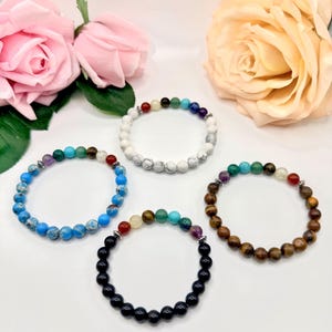 May include: Four beaded bracelets with various coloured stones. One bracelet is blue, another is white, a third is black, and the last is brown. The bracelets are displayed with pink and yellow roses in the background.