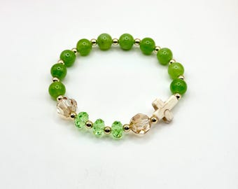 May include: A beaded bracelet featuring round, light green beads interspersed with small gold-colored beads. The bracelet includes faceted crystal beads in light green and tan, and a small, off-white cross charm. The bracelet is on a white background.