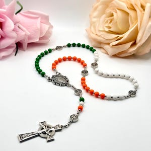 May include: A rosary featuring green, orange, and white beads arranged in a sequence, connected by silver-toned metal links. The rosary includes a silver-toned crucifix and a central medal depicting a figure, accented with small silver-toned spacer beads.