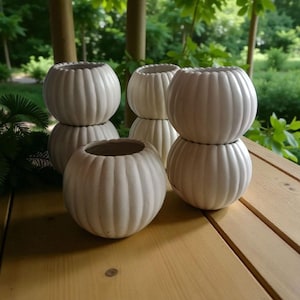 May include: A collection of six off-white, round, ribbed ceramic planters. The planters are stacked in pairs and arranged on a wooden surface. The background features green foliage and wooden posts, suggesting an outdoor setting. The planters have a pumpkin-like shape.