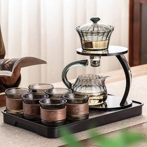 May include: A modern tea set with a glass teapot, a serving stand, and six matching glasses. The teapot and glasses have a ribbed design and are displayed on a black tray. The glasses have brown leather-like wraps.