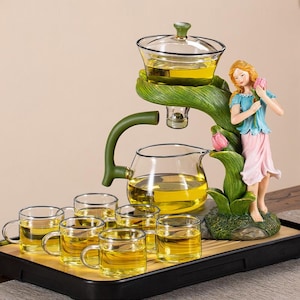 Puede incluir: Un juego de té con una tetera de vidrio, un infusor y seis pequeñas tazas de vidrio llenas de té. Un soporte decorativo en forma de hoja verde sostiene el infusor y una figura de mujer. El juego está sobre una bandeja negra.