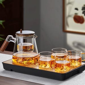 Puede incluir: Un juego de té de vidrio transparente con asa y tapa de madera. El juego incluye una tetera con infusor, cuatro tazas y una bandeja de madera. El té es de color ámbar.