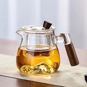 Puede incluir: Una tetera de vidrio transparente con asa y tapa de madera. La tetera está llena de té de color ámbar y la base tiene un diseño similar a una montaña. El asa y la tapa son de madera oscura.