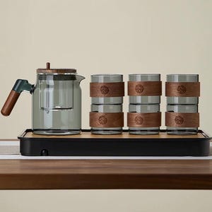 Puede incluir: Un juego de té con una tetera de vidrio con asa y tapa de madera, y seis vasos a juego. Los vasos tienen bandas de madera con un emblema decorativo. El juego se exhibe en una bandeja negra.