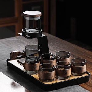 Può includere: Un set da tè con una teiera in vetro e sei bicchieri piccoli, ciascuno con una custodia in legno. Il set include una brocca da portata con manico in legno e un vassoio. La teiera ha un coperchio in legno e poggia su un supporto in metallo nero.