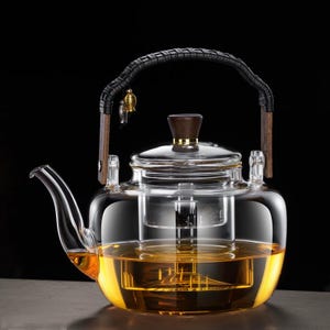 Puede incluir: Una tetera de vidrio transparente con asa y tapa de madera. La tetera está llena de un líquido de color dorado, probablemente té. El asa está envuelta en material negro. Un pequeño adorno dorado cuelga del asa. La tetera está diseñada para preparar té.