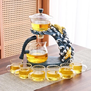 May include: A glass tea set with a decorative stand featuring a wave design and golden dolphin accents. The set includes a brewing pot, a serving carafe, and six small teacups, all filled with golden tea. The stand is blue and white.