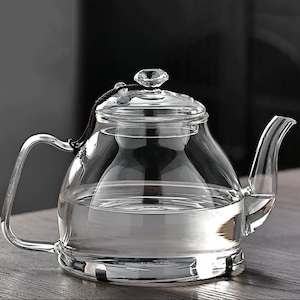 May include: A clear glass teapot with a curved handle and spout. The teapot has a glass lid with a crystal-like knob. The teapot is filled with water, suggesting it is ready for brewing tea. The design is simple and elegant.