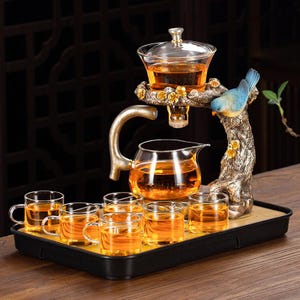 Può includere: Servizio da tè decorativo con teiera in vetro, tazze e supporto ornamentale. Il supporto presenta un uccello blu e dettagli floreali. Il tè è ambrato e il set è su un vassoio nero.