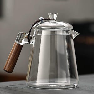 Op de afbeelding: Een heldere glazen theepot met een houten handvat en een glazen deksel. De theepot heeft een tuit om uit te schenken en een klein decoratief element op het deksel. Het handvat is gemaakt van donker hout en is bevestigd aan de pot met een zilverkleurig metalen stuk.