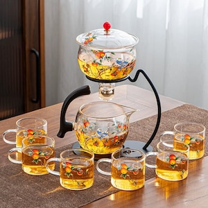 Può includere: Un set da tè in vetro trasparente con un design decorativo di arance e uccelli. Il set comprende una teiera, una brocca e sei tazze con manico. Il set da tè è su un tavolo marrone.