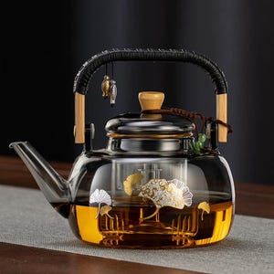 Puede incluir: Una tetera de vidrio transparente con un asa negra y detalles de madera. La tetera presenta un diseño de hojas doradas y plateadas y está llena de líquido color ámbar. Un pequeño amuleto cuelga del asa.